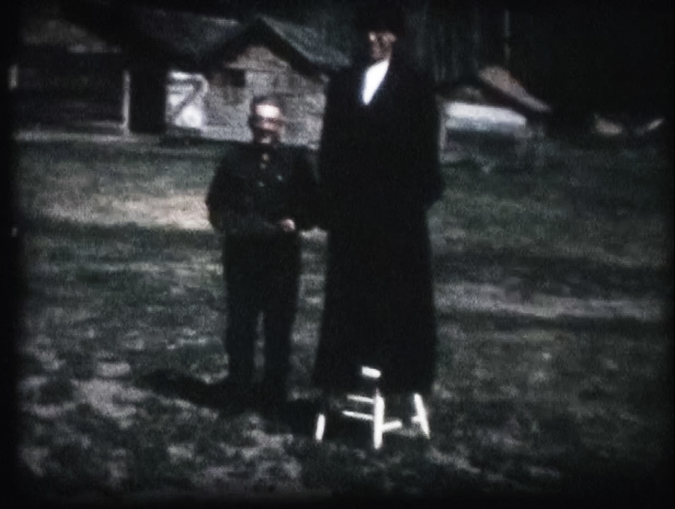 Capture d’image d’un film Super 8 d’Alcide Short filmé chez Georgiana Beaupré et Joe Short à Park Valley, SK. Sur la photo, un homme porte le manteau énorme d’Edouard et l’autre tient sa grande chaussure.